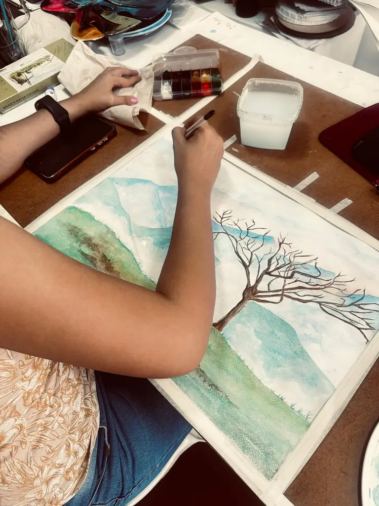 Manos de una estudiante pintando en acuarela un árbol y un paisaje de montañas.